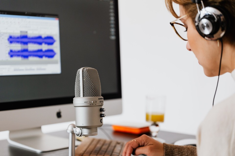 A person wearing headphones and speaking into a microphone, with a computer screen displaying audio editing software in the background