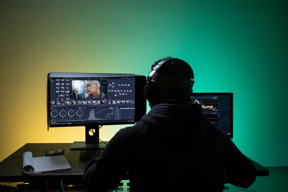 A person wearing headphones sits in front of dual monitors, working on video editing software, with a gradient yellow to green background