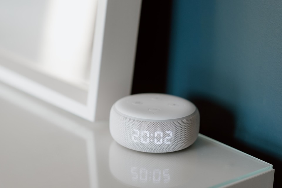 A white smart speaker with an LED display showing the time 20:02, placed on a glass surface