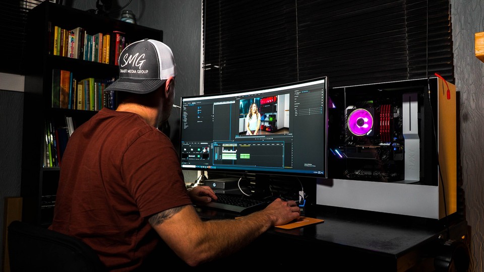 A person working on video editing software on a dual-monitor setup in a home office with a bookshelf in the background