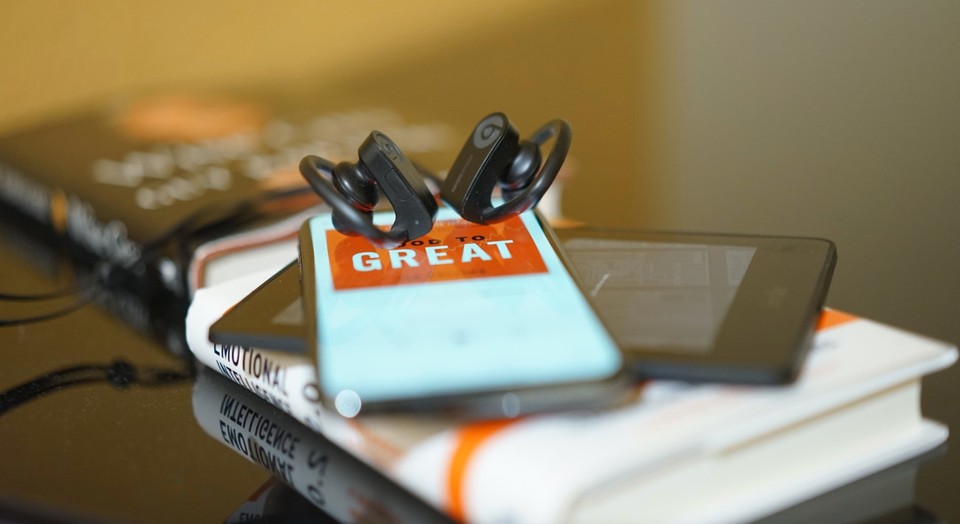 A pair of headphones resting on a smartphone displaying a book titled Good to Great, with other books in the background