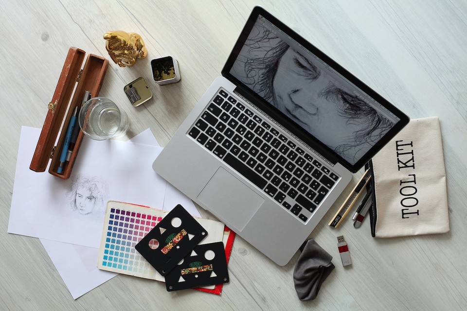 A top-down view of a workspace with a laptop displaying a sketch, drawing tools, a color palette, and various art supplies