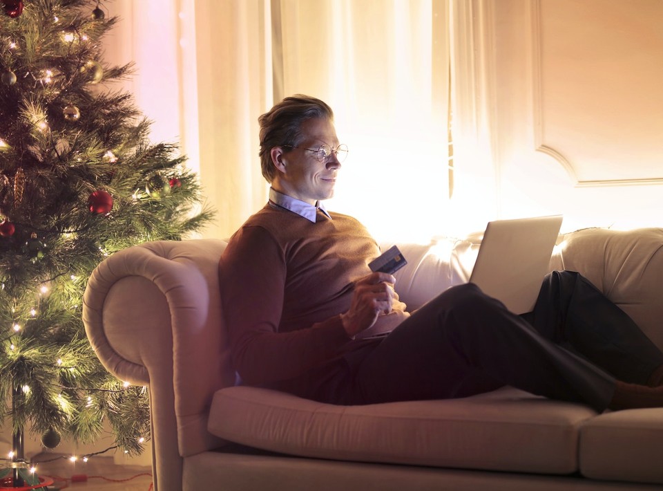 Man holding a credit card while shopping online on a laptop, sitting on a sofa near a Christmas tree