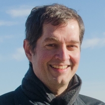 A man with short dark hair smiling while wearing a black jacket, set against a blue sky background